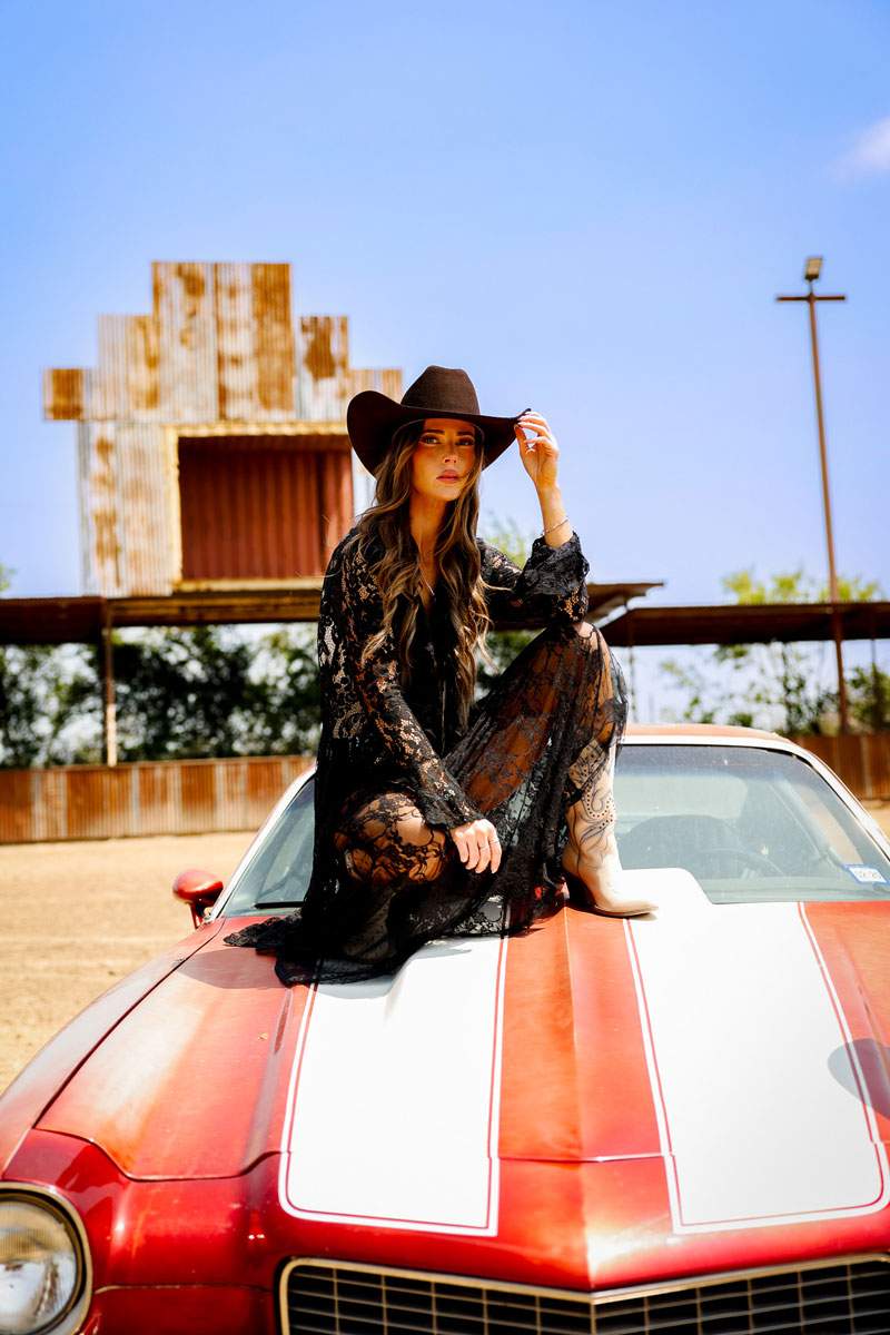 Woman in a black hat and black lace dress seated on a striped vintage Camaro at a rustic ranch — Western fashion portrait by BK Bennett